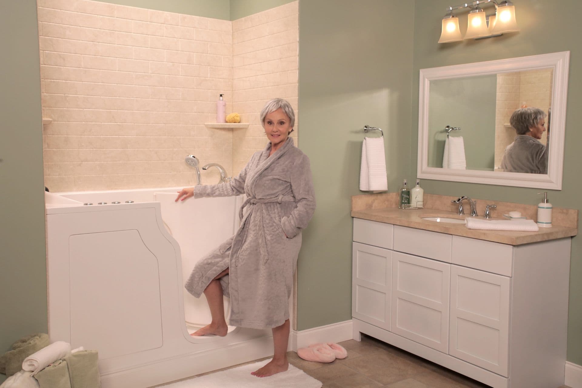 Senior woman demonstrates walk-in bathtub with door open in accessible bathroom featuring vanity sink and framed mirror.